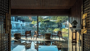 Ein Zimmer mit einem großen Fenster, Holzboden und Blick auf einen Garten mit Bäumen und einem Teich.