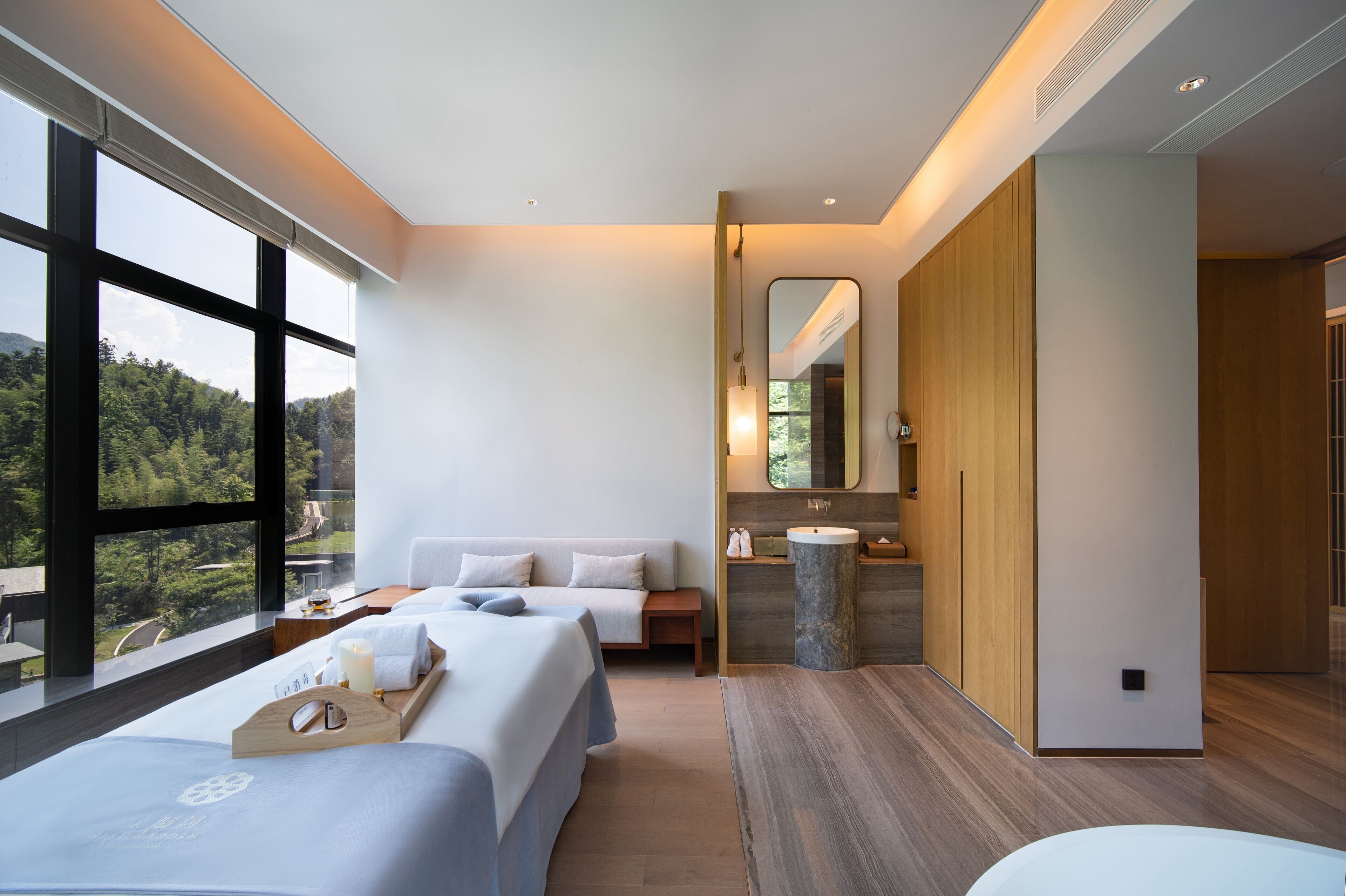 Quarto de hotel moderno com uma janela grande, duas camas, piso de madeira, banheiro com espelho e pia, e vista para o exterior.