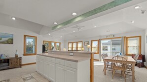 A spacious kitchen with white cabinetry, a central island, and a dining area with a wooden table and chairs. - 1750 - Cool Winds, Corolla Beach House: Private Pool & Hot Tub (Corolla)