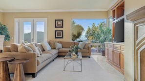 Una sala de estar con un sofá color beige, una mesa de centro y un televisor. Hay dos ventanas con vistas a árboles y colinas.