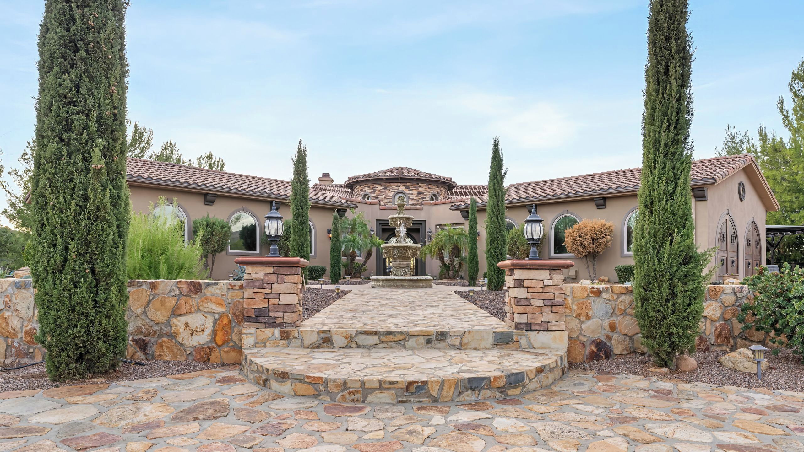 A house with a stone pathway leading to a central fountain, surrounded by tall cypress trees and a stone wall.