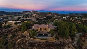A large, single-story house with a spacious patio and a swimming pool, surrounded by a fenced property and dense trees.