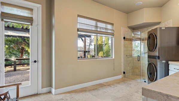 A kitchen with a built-in washer and dryer, a glass shower enclosure, and a door leading to an outdoor area with a bench and trees. - Hilltop Estate w/ Pool, Hot Tub, Fire Pit, Views Cleveland Vista by Avantstay (Murrieta)