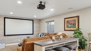 A living room with a brown leather sofa, a wooden coffee table with two stools, a projector screen, and a framed picture on the wall. - The Fritz | Pool + Theater + Games | Sleeps 14 (Glendale)