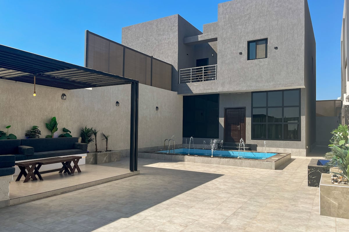 A modern outdoor area with a pool, a seating area with a sofa and table, and a pergola.