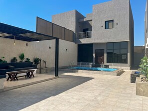 Un espacio exterior moderno con piscina, zona de estar con sofá y mesa, y una pérgola.