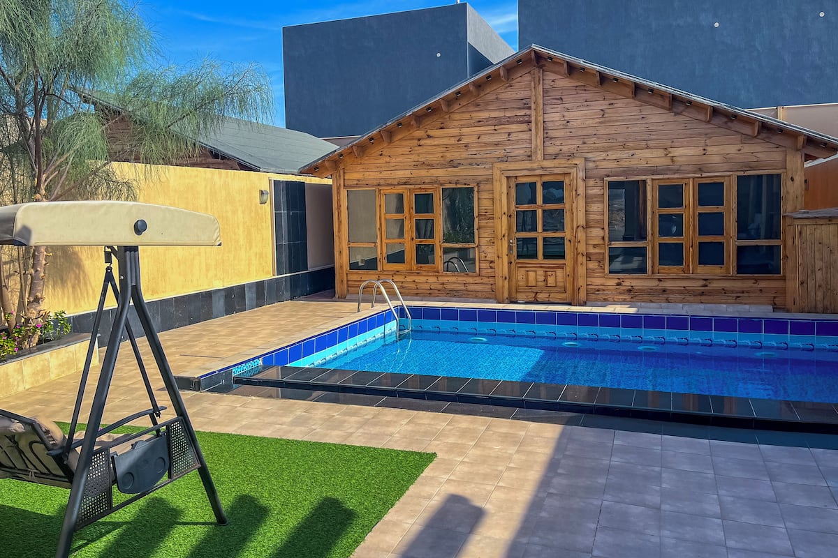 A wooden cabin with a pool, surrounded by a patio area with a swing set.