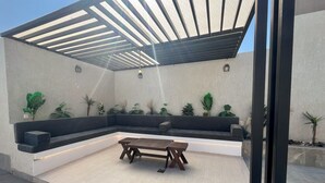 Zona d'estar exterior moderna amb una cadira incorporada i taula de fusta, envoltada d'arbres en testos i una estructura de pergola.