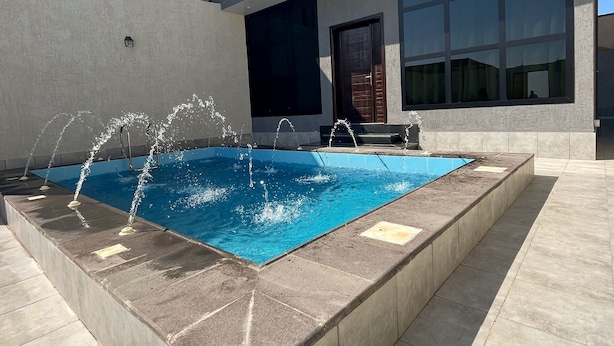 Zona de piscina exterior moderna amb una piscina rectangular, envoltat per un solàrium de pedra, i un edifici amb finestres grans i un balcó.