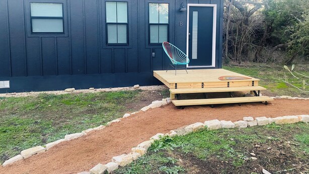 Une petite maison de couleur sombre, avec une porte, des fenêtres et une chaise à l’extérieur.