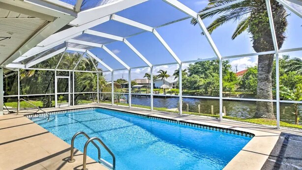 A pool area with a glass enclosure, clear blue water, and a view of a lake and palm trees. - Tropical Getaway! Gulf access with heated pool! (Cape Coral)