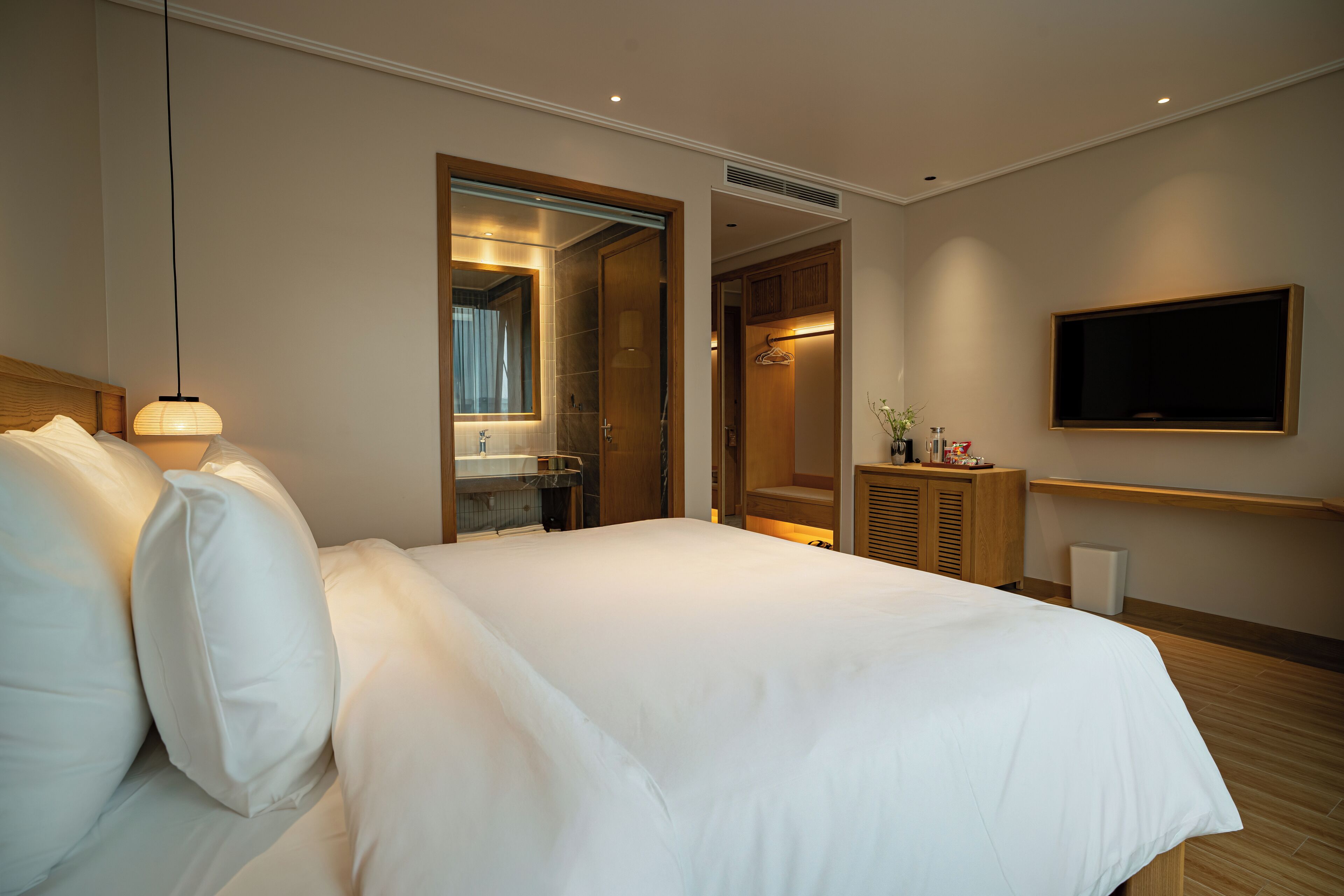 Habitación de hotel con una cama grande, una lámpara de noche, un espejo, un televisor y una mesita de noche con artículos de tocador.