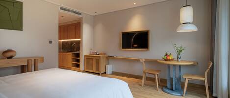 Habitación de hotel moderna con una cama grande, una mesa de comedor con sillas, una pequeña cocina y un televisor montado en la pared.
