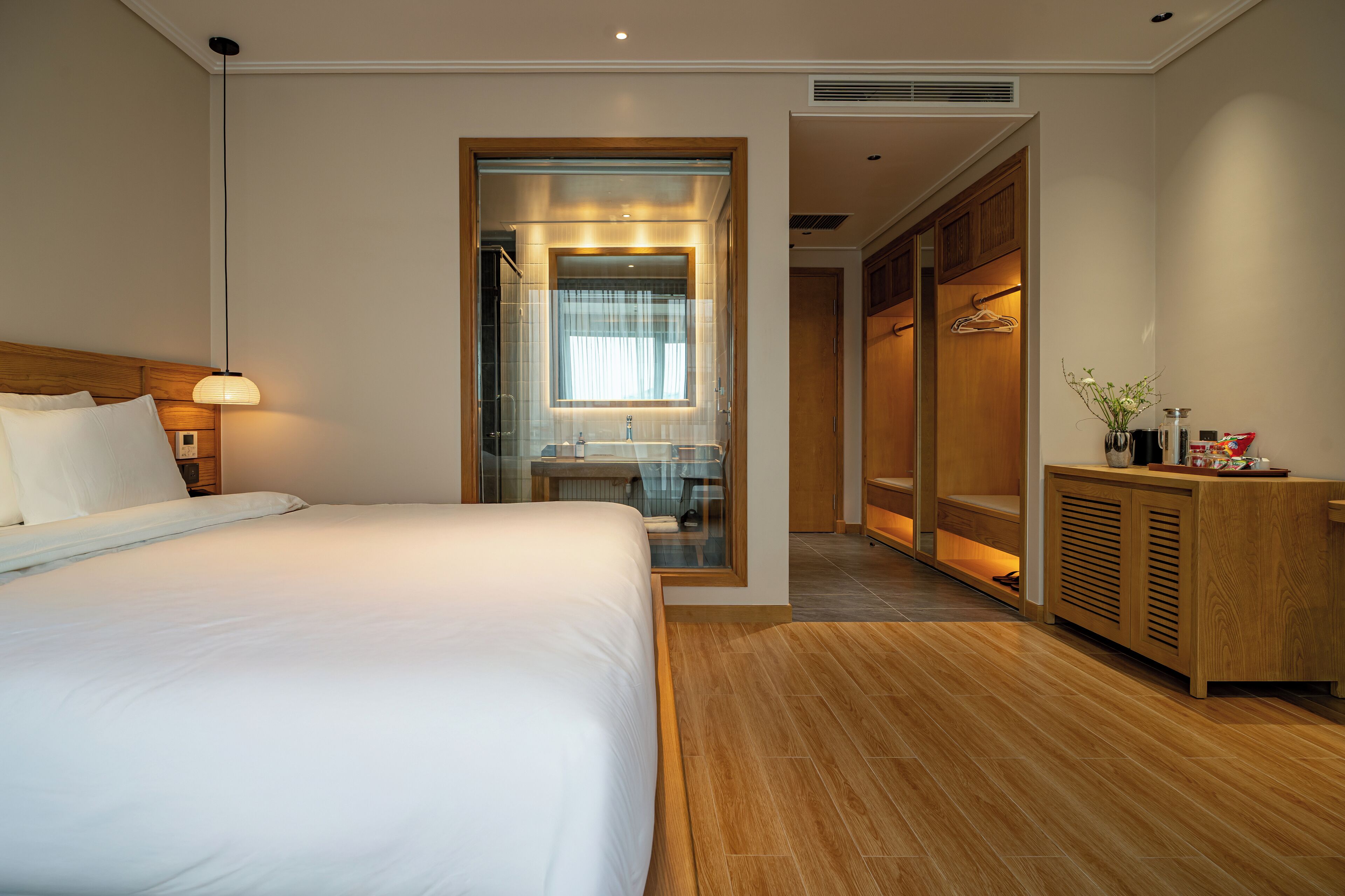 Una habitación de hotel moderna con una cama grande, un armario de madera, un tocador con espejo y un mueble pequeño con artículos de aseo.