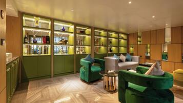 A modern interior with green and wooden accents, featuring a seating area with green upholstered chairs and a white sofa, and a bar area with glass display shelves.