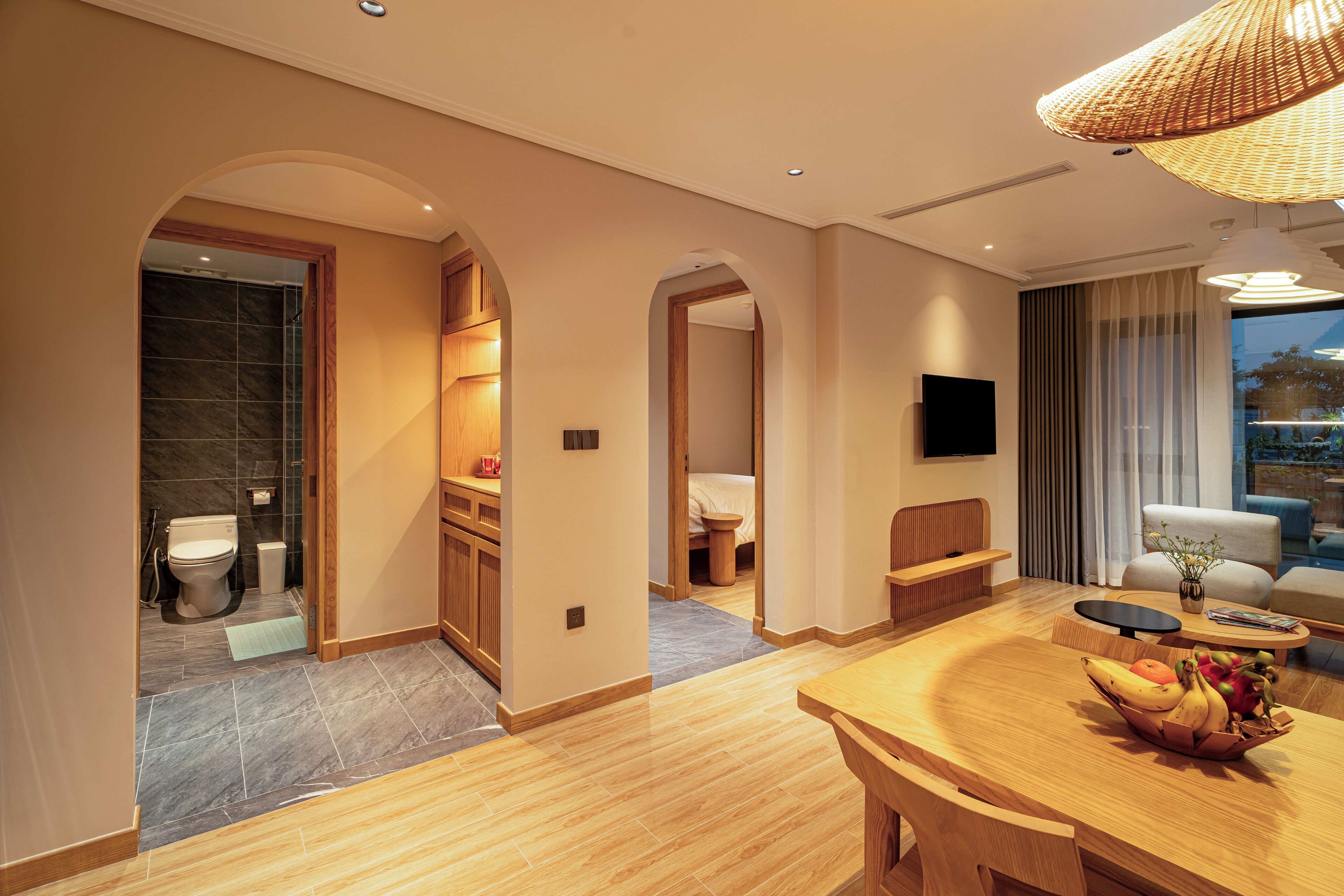 A modern hotel room with a wooden dining table, a TV mounted on the wall, and a view of the outdoors through the window.