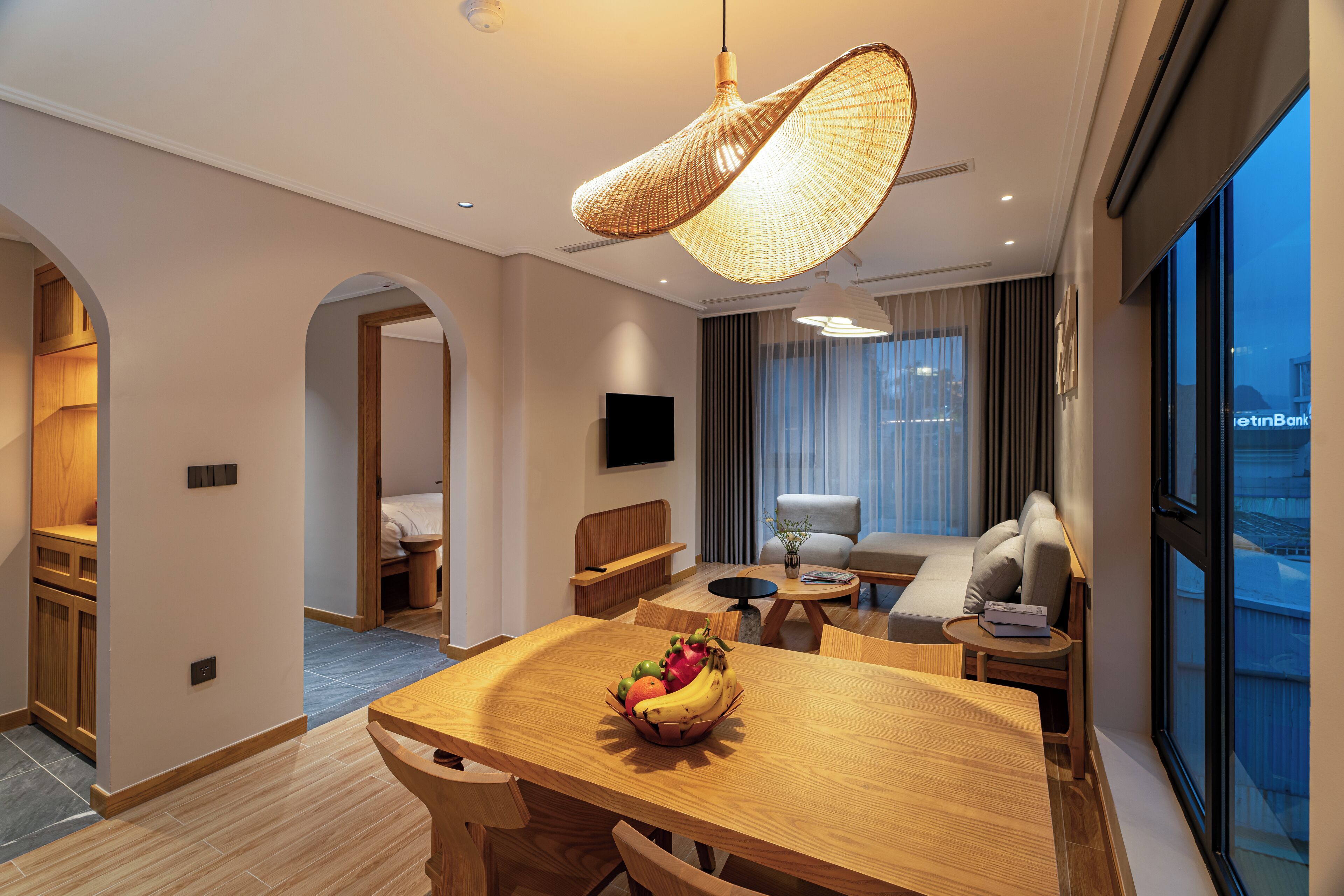 A modern hotel room with a wooden dining table, a fruit bowl, a sofa, and a large window offering a city view.