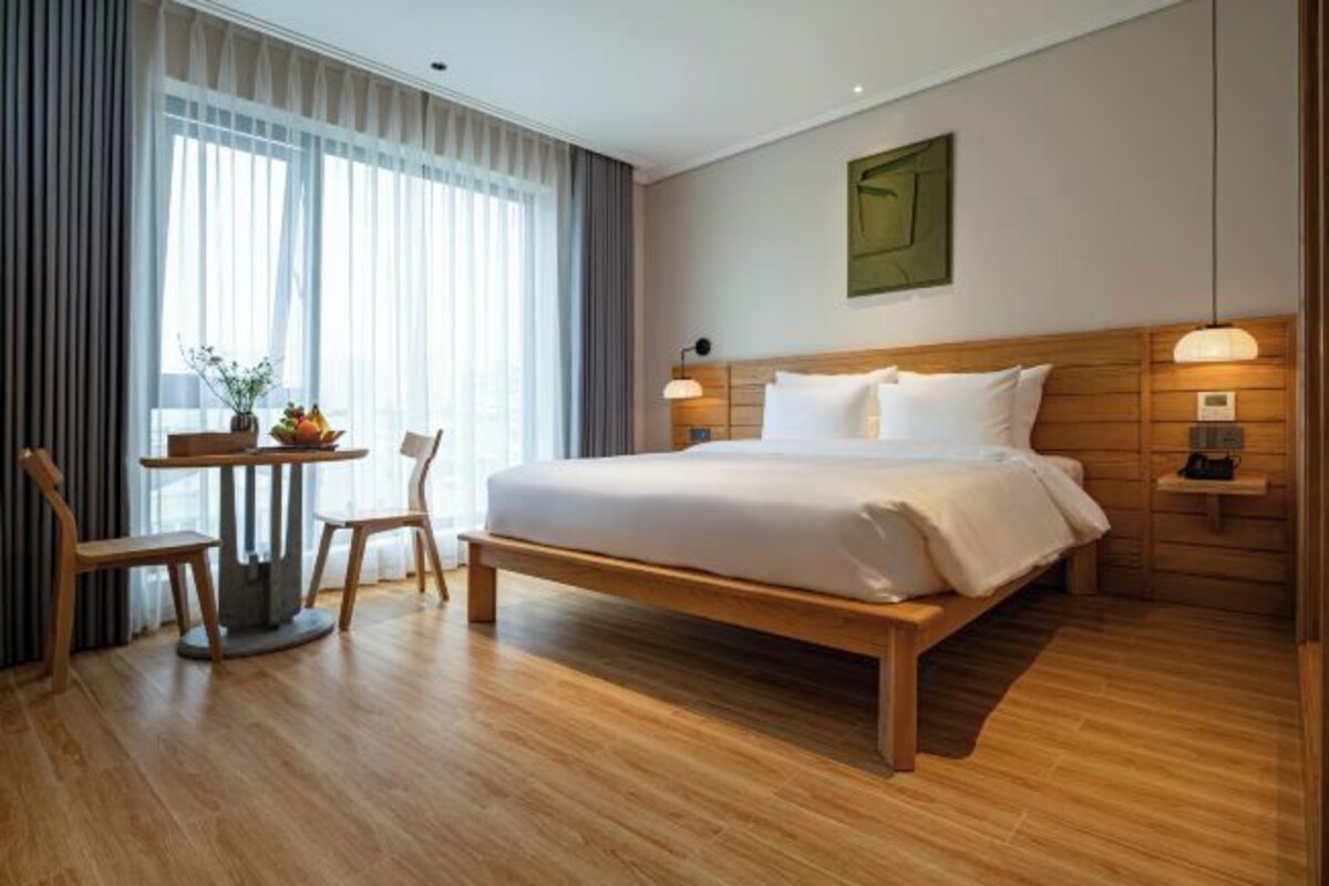 Habitación de hotel con cama de madera, una pequeña mesa redonda con sillas, un frutero y vistas a la ciudad a través de ventanas amplias.