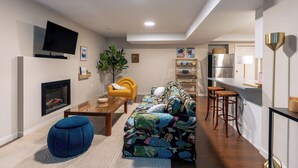 Ein modernes Wohnzimmer mit einer Couch, einem Couchtisch, einem blauen Hocker, einem Fernseher und einem Kamin.