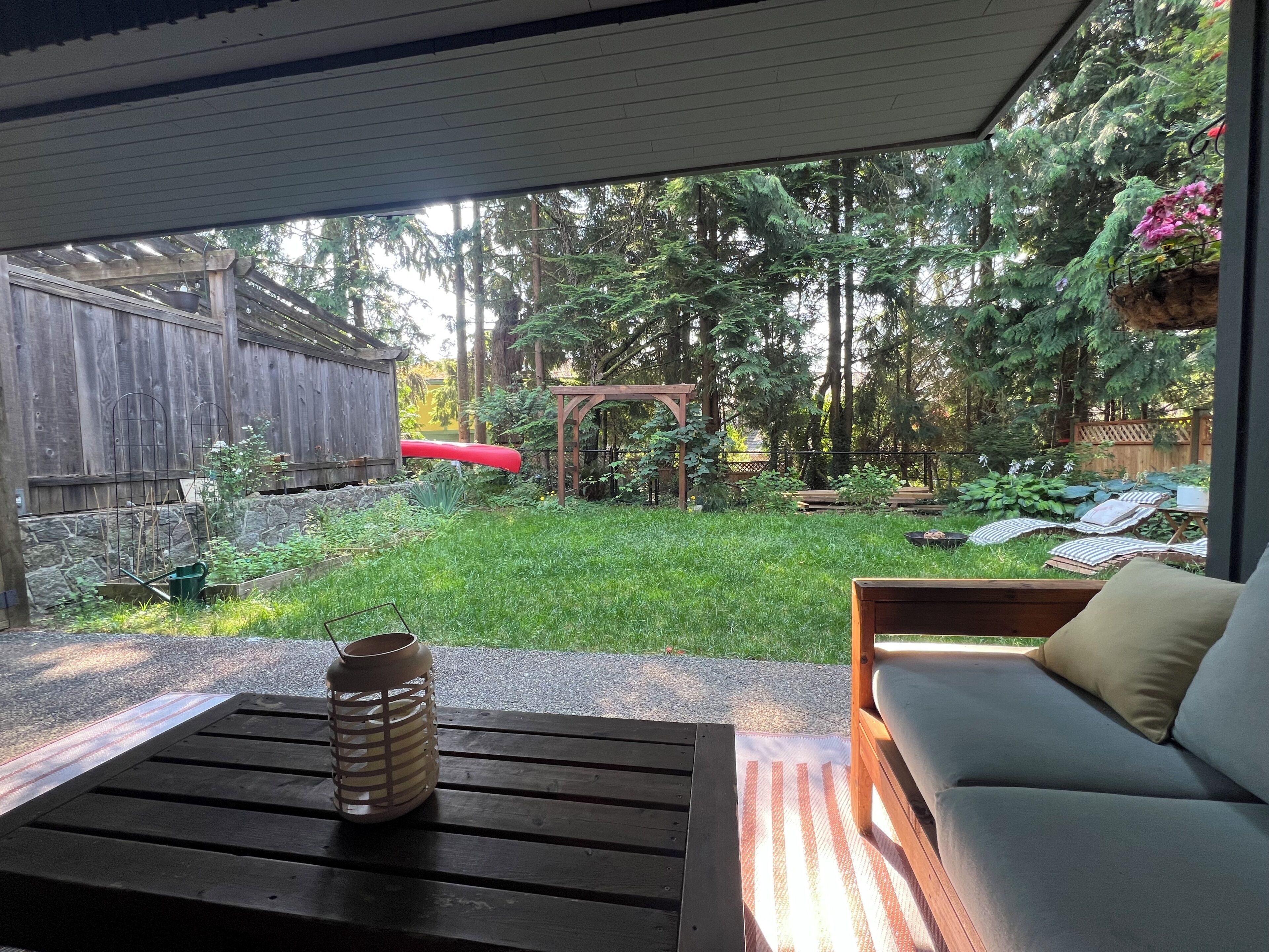 A patio with a wooden table and a cushioned seating area, overlooking a backyard with a wooden fence, a red canoe, and a wooden pergola.