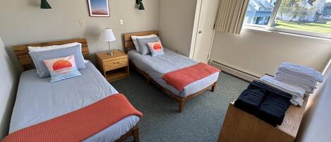 A hotel room with two single beds, a wooden nightstand with a lamp, a window with curtains, and a stack of towels on a wooden dresser.