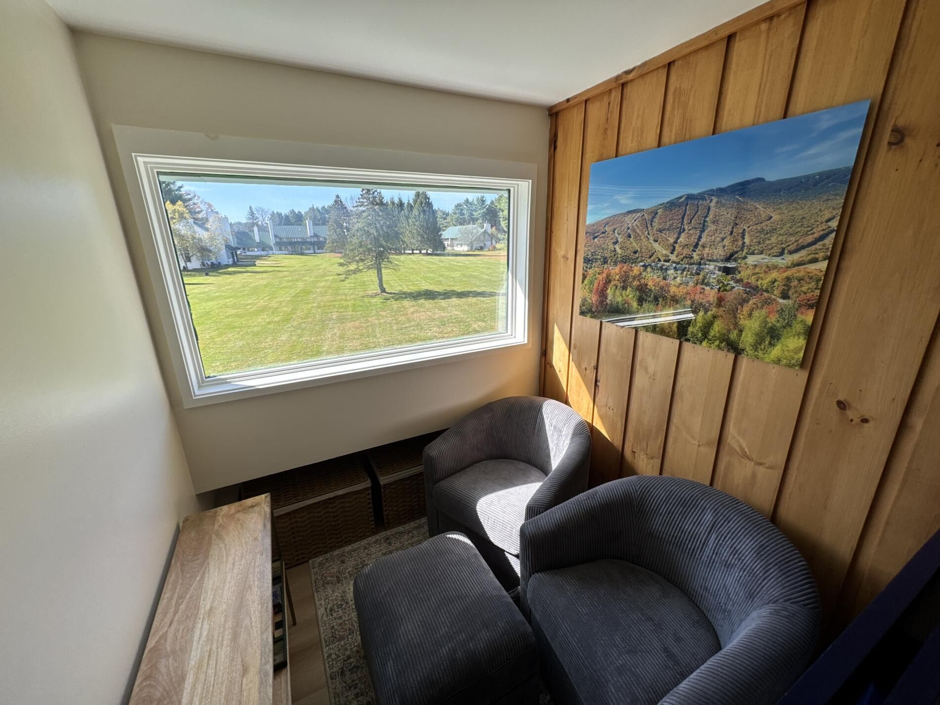 A room with a large window offering a view of a grassy area and trees, a wooden wall with a framed landscape painting, and two upholstered chairs.