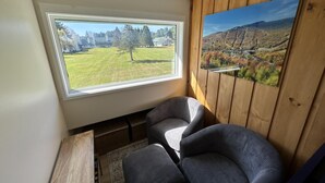 Ein Zimmer mit großem Fenster und Blick auf eine Wiese und Bäume, eine Holzwand mit einem gerahmten Landschaftsbild sowie zwei Polsterstühle.