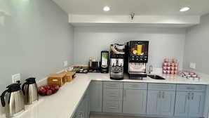 Una cocina moderna con cafetera integrada, dispensador de agua y pileta. Se observan muebles de cocina, una mesada y algunos objetos como manzanas y una cesta.