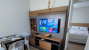 Ein modernes Hotelzimmer mit einem Holz-Kopfteil, einem flachbildfernseher an der Wand, einem Esstisch mit Stühlen, einem Sofa und einem Bett mit weißer Bettwäsche.