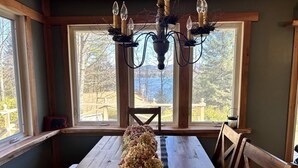 A dining area with a wooden table, chairs, and a chandelier. Large windows provide a view of the outdoors. - Adirondack Cabin (Schroon Lake)