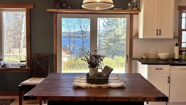 A wooden dining table with a hanging light fixture, a view of a lake through large windows, and a kitchen area with white cabinets. - Adirondack Cabin (Schroon Lake)