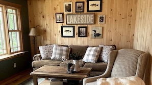 A cozy living room with a wooden ceiling, a large window, and a fireplace. There are framed pictures on the wall and a coffee table in the center. - Adirondack Cabin (Schroon Lake)