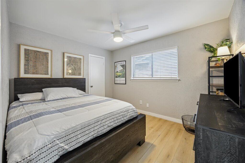 Une chambre à coucher avec un lit, un ventilateur de plafond, une fenêtre avec des stores et un téléviseur posé sur un meuble.