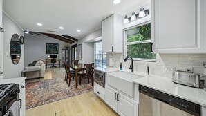 A modern kitchen with white cabinetry, a built-in dishwasher, and a toaster. There is a dining area with a wooden table and chairs, and a large window providing natural light. - NEW Charming 3BR 2BA w/Modern Fireplace by Domain @ Q2 Stadium (Austin)