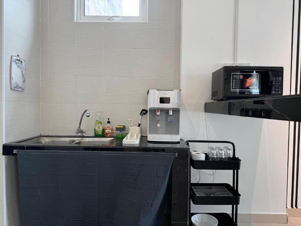A small kitchenette with a coffee maker, a microwave, and a water dispenser. There is a sink with a faucet, and a shelf with glasses and a small container.