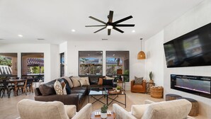 Un salón amplio con un moderno ventilador de techo, un televisor de pantalla plana grande montado en la pared y una zona de estar confortable con sofá y sillones.