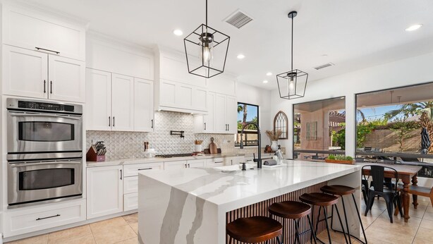 Una cocina moderna con armarios blancos, electrodomésticos de acero inoxidable, una isla grande con taburetes, iluminación colgante y vistas al exterior.