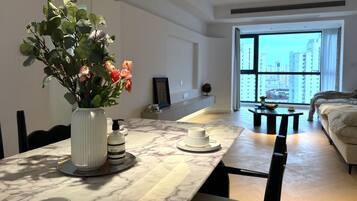 A modern living room with a dining area, featuring a marble table, a vase with flowers, and a large window offering a city view.
