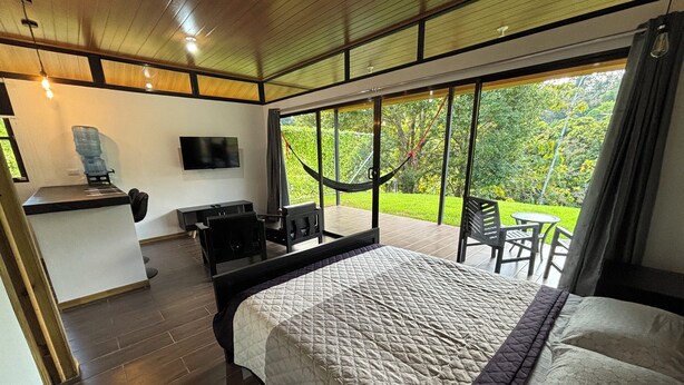 Un dormitorio con una cama grande, un escritorio, un televisor y vistas a un jardín a través de amplios ventanales.