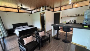 Una habitación compacta con cama, un pequeño rincón de descanso, kitchenette y ventilador de techo.