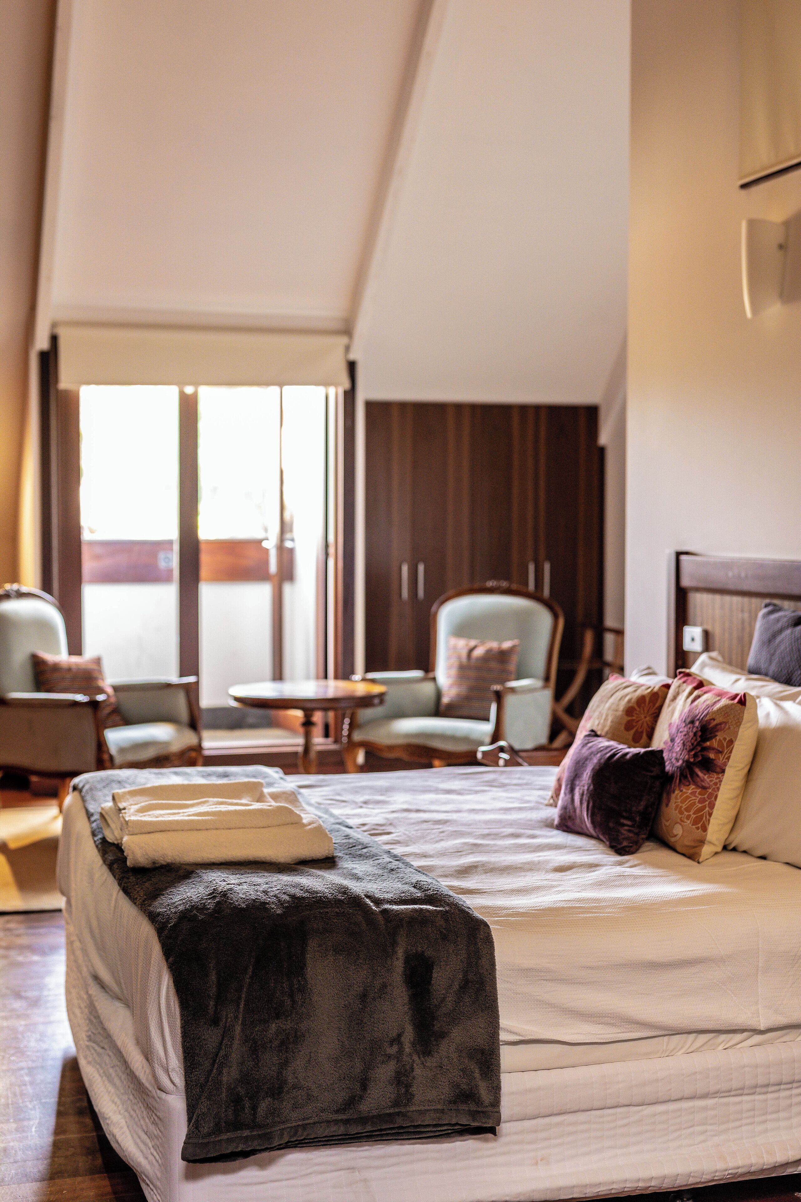 A hotel room with a bed, a chair, a small table, and a view of the outside.