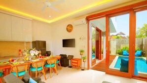A modern living room with a dining area, a TV mounted on the wall, and a view of a swimming pool through glass doors. - Luxury Villa Noam 2BR Nusa Dua (Nusa Dua)