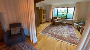 Un salon spacieux avec un grand tapis, un plancher en bois, un fauteuil sombre, une fenêtre donnant sur la verdure et un meuble encastré.