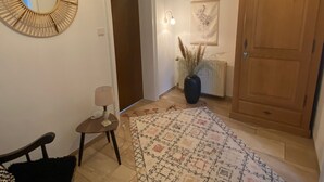Un couloir avec un miroir, une porte, une lampe, un radiateur, un vase avec des plantes, un tapis à motifs et un meuble en bois.
