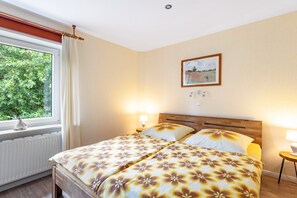 A bedroom with a bed, two bedside lamps, a window with curtains, a radiator, and a framed picture on the wall.