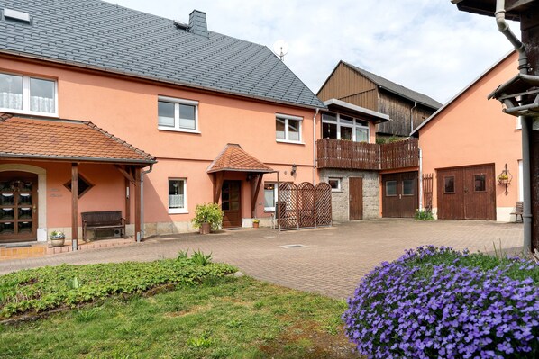 A courtyard with a brick pathway, a wooden fence, and a building with a sloped roof and a balcony. - Holiday Apartment '2' with Shared Terrace, Shared Garden and Wi-Fi (Hohnstein)