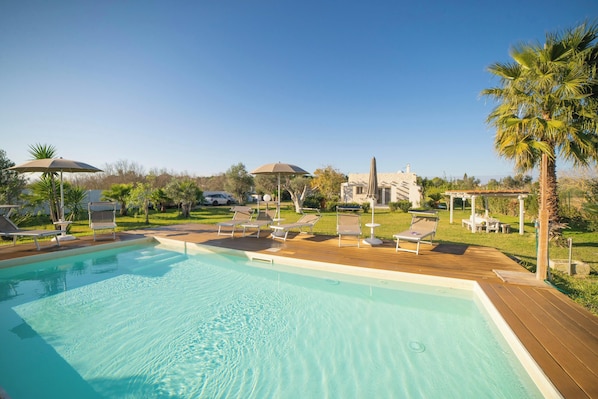 A swimming pool with lounge chairs and umbrellas, surrounded by greenery and a stone building.