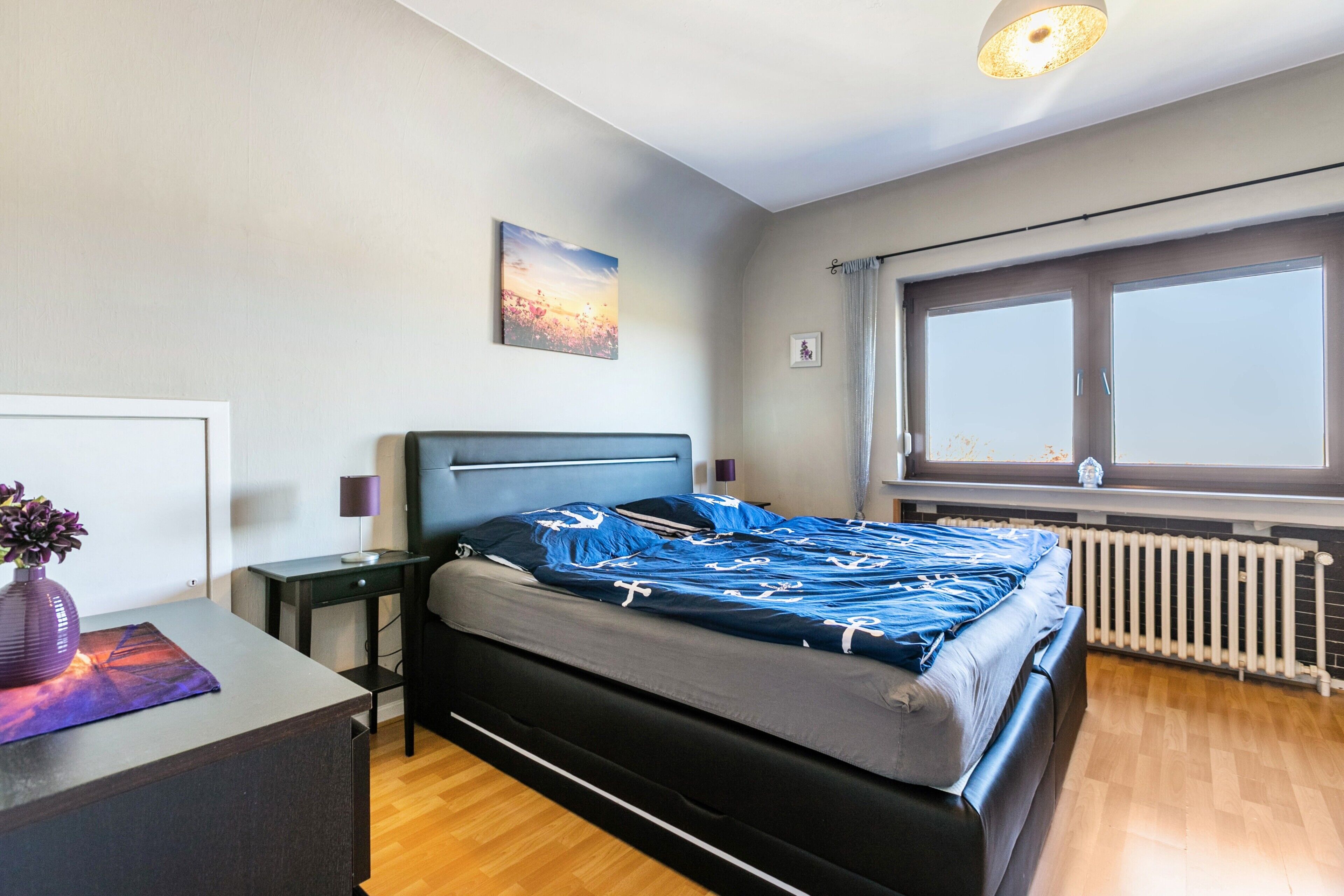 A modern bedroom with a bed, bedside tables, a nightstand, a window with curtains, a radiator, and a wall-mounted picture.