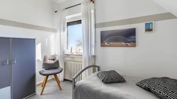 Un dormitorio con cama, una silla, armario, ventana con cortinas y un cuadro en la pared.