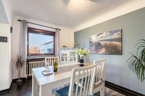 A dining area with a white table, chairs, a bottle of wine, a vase with flowers, and a coastal landscape painting on the wall.